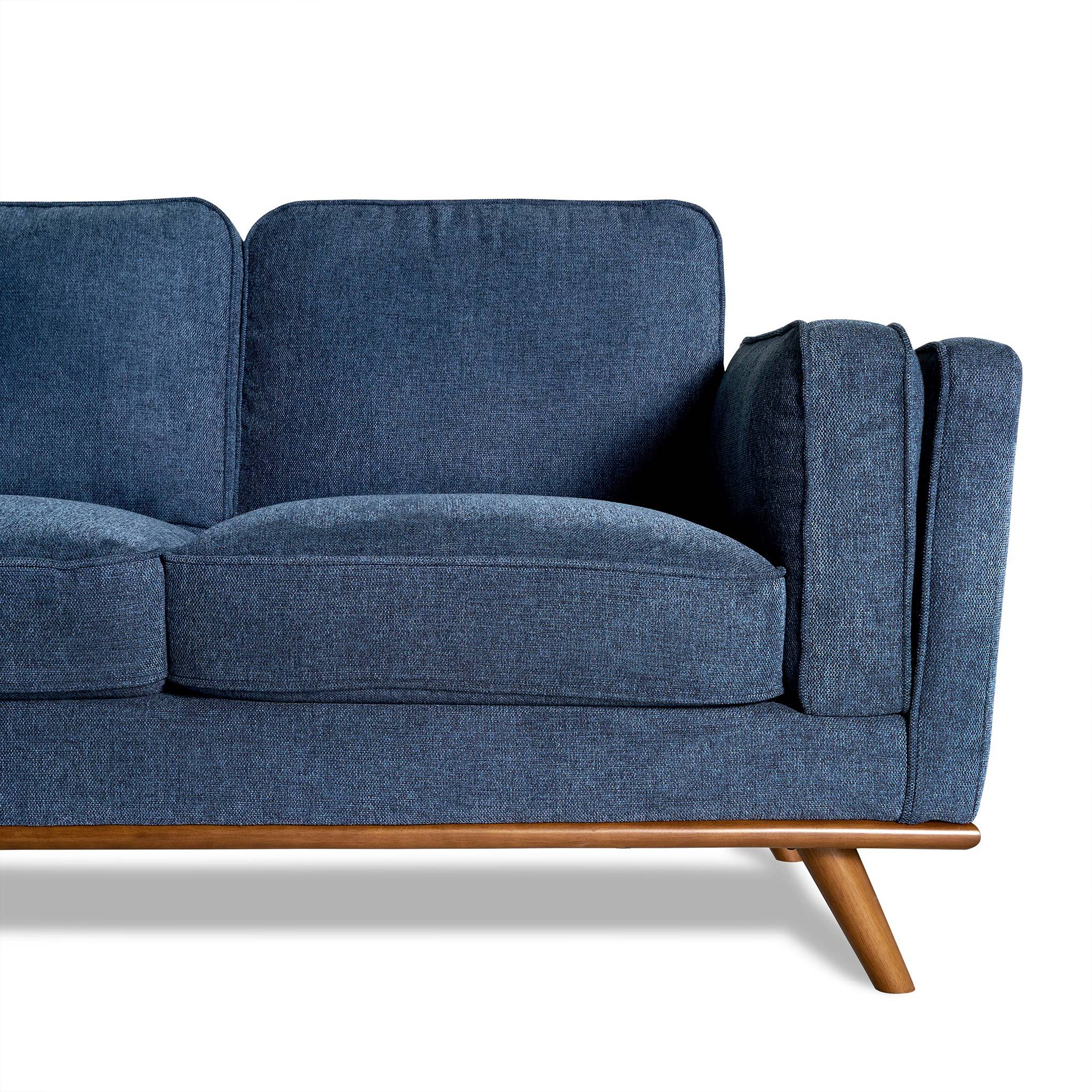The Maddox Sectional, with its blue fabric upholstery and mid-century modern style, features wooden legs and armrests. Set against a white background, its plush cushions bring elegance to any living room. The focus is on the right half of this minimalist design.