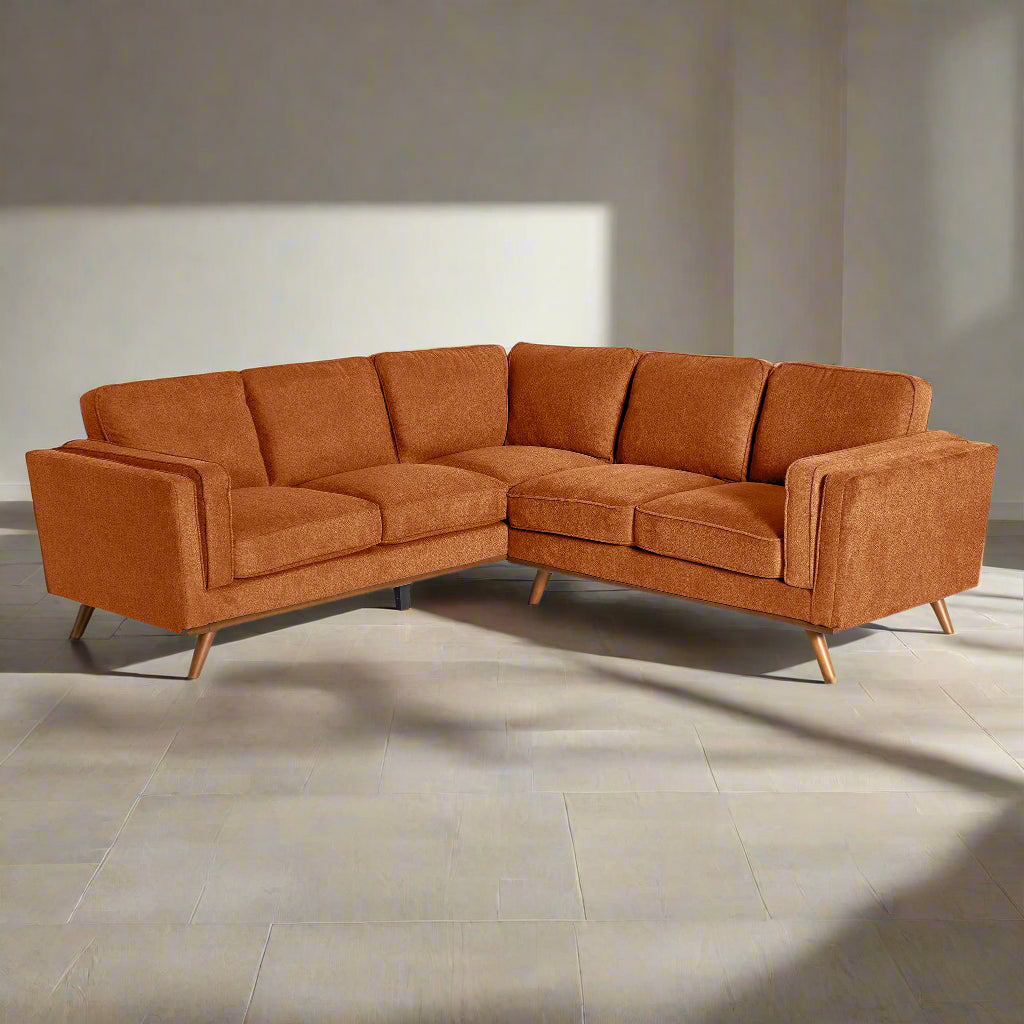 The Maddox is a mid-century modern L-shaped sofa with burnt orange upholstery, plush cushions, and sleek tapered wooden legs against a white background.