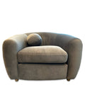 The Sculpture Chair is a plush, gray armchair with sculptural curves and a rounded design. It boasts luxurious upholstery, includes a cylindrical pillow on one side, and features short wooden legs that enhance its minimalist style, set against a plain white background.